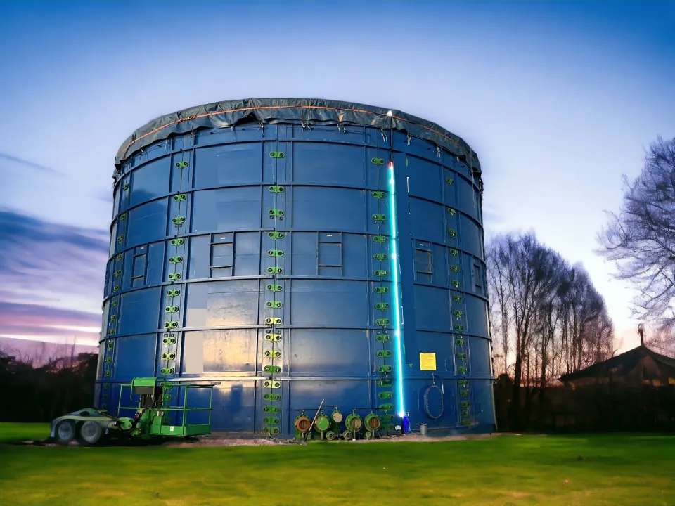 A large blue bolted-steel storage tank at dusk with a DCon SHIELD LED level indicator glowing on its side
